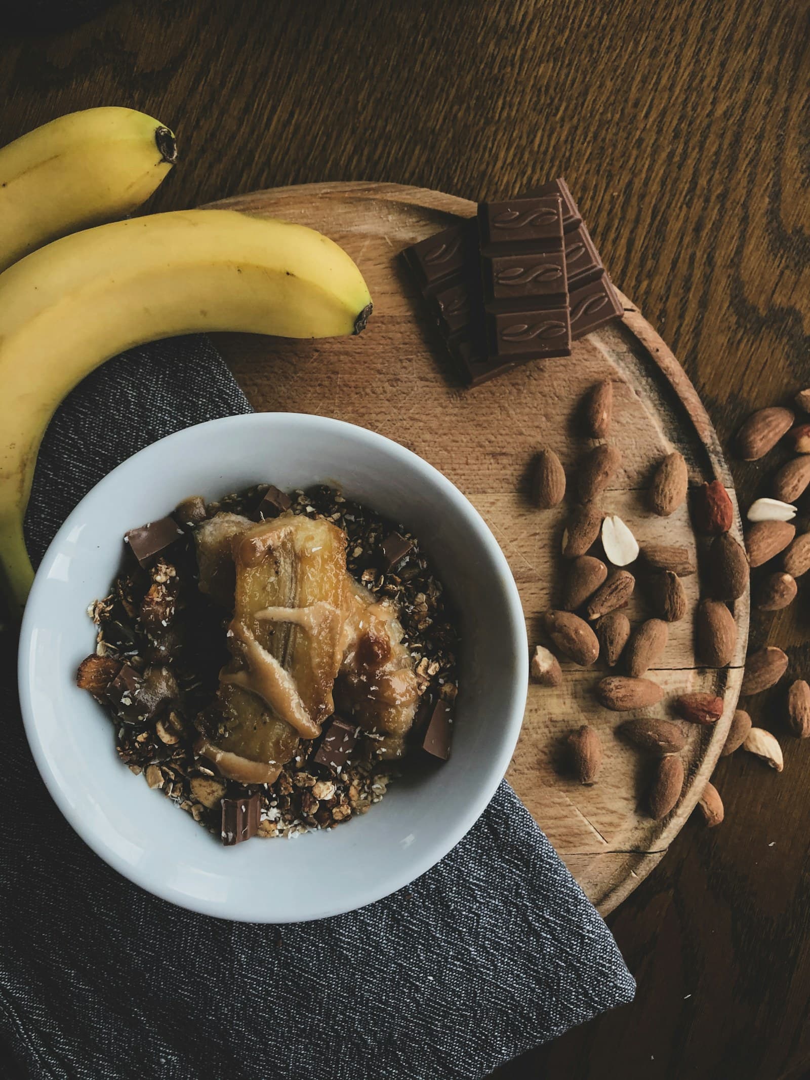 A bowl of oats and banana beside whole bananas and a pile of almonds on a wooden board